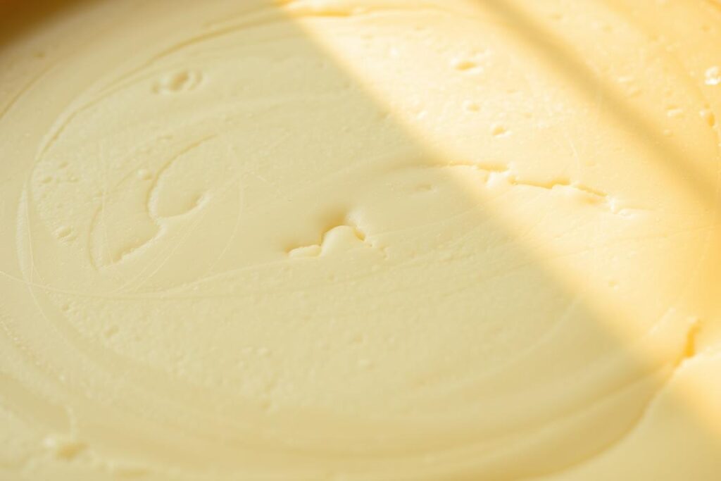 A close-up view of a perfect cheesecake surface, showcasing a smooth and creamy texture with a subtle golden-brown hue. The surface is free of any cracks or blemishes, reflecting an even and well-baked interior. Delicate swirls and patterns are visible, hinting at the care and attention taken during the preparation process. Soft, natural lighting casts a warm glow, accentuating the luxurious appearance of the dessert. The image captures the essence of a professionally crafted cheesecake, inspiring the viewer to imagine the decadent flavor and satisfying texture.
